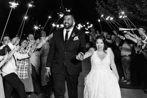 Black and White photo of newly married man and woman exiting wedding with guests holding long wedding sparklers