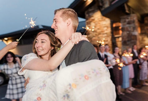 Groom carrying bride out of wedding venue with guests holding long wedding sparklers