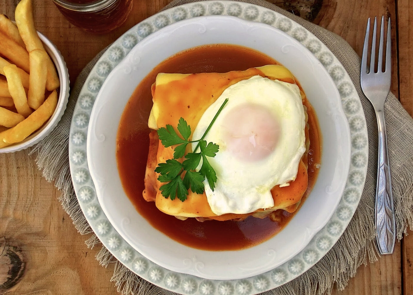 Auf einem weißen Teller liegt eine köstliche Francesinha, eine portugiesische Delikatesse. Das Gericht besteht aus einem saftigen Toast mit zartem Fleisch und Würstchen, überzogen mit geschmolzenem Käse und einer reichhaltigen Sauce. Ein perfekt gebratenes Spiegelei thront auf der köstlichen Kreation. Dazu serviert werden goldene Pommes-frîtes, die das Bild eines verlockenden kulinarischen Erlebnisses abrunden.