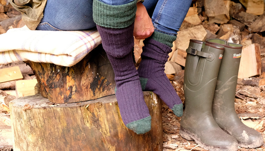 closeup of person sitting on a log beside a pair of wellies, wearing warm, winter Corgi socks