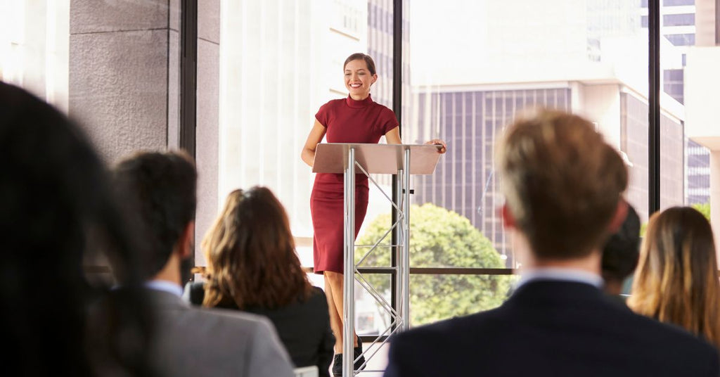 A woman speaking in front of an audience using a metal lectern