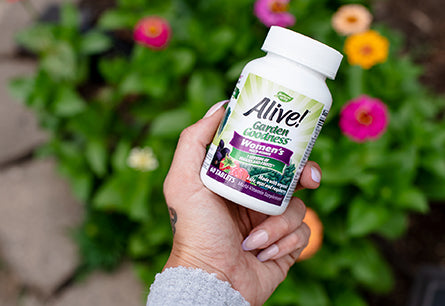 A hand holding a bottle of Alive Garden Goodness for Women with flowers in the background.