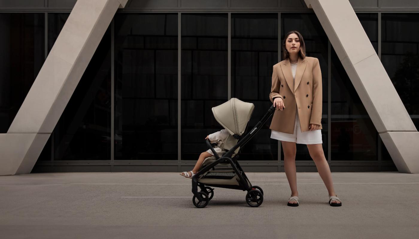 A lifestyle photo featuring a woman in a beige blazer and white dress, standing next to the Redsbaby AERON stroller in an urban setting. The stroller seat is reclined with a child resting inside, and the canopy is extended halfway. The modern architecture of the surroundings emphasizes the stroller's stylish design.
