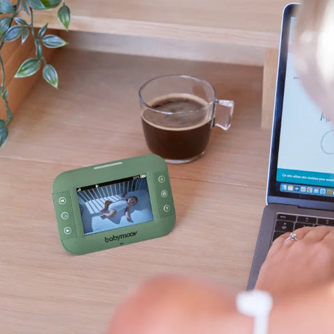 Φωτογραφία τρόπου ζωής ενός σκηνικού σε ένα γραφείο στο σπίτι. Η οθόνη της κύριας κάμερας babymoov yoo τοποθετείται στο πλάι ενός φορητού υπολογιστή, μπροστά από ένα μισό γεμάτο φλιτζάνι καφέ. Στην οθόνη φαίνεται το μωρό να παίζει μέσα στο δωμάτιό του.