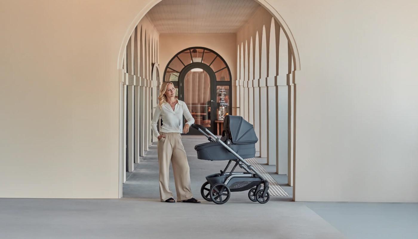 A woman stands in a spacious, modern hallway with arched doorways, pausing to glance back at the camera while holding onto a Redsbaby NUVO stroller. The stroller, featuring a bassinet in a sophisticated slate blue color, complements the woman's elegant cream blouse and beige trousers. The architectural environment, bathed in soft natural light, accentuates the stroller's luxurious design.