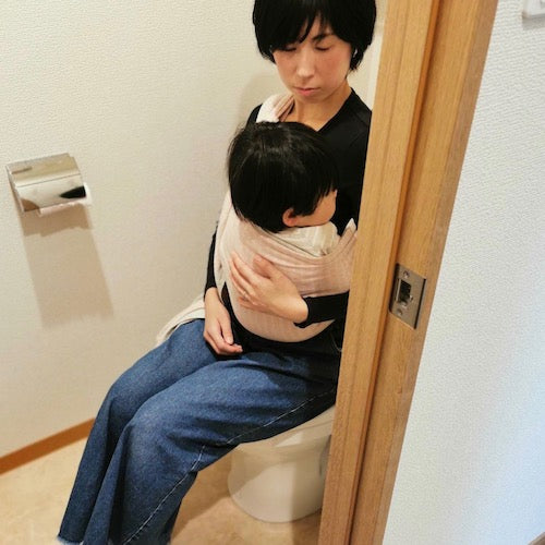 スリングで赤ちゃんを抱っこしてトイレに座るお母さん