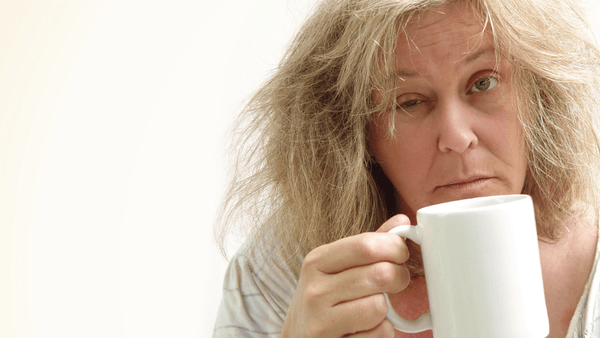 Eine geränderte Frau guckt Müde in die Kamera während sie eine Tasse Kaffee als Wachmacher trinkt.