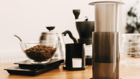 Eine AeroPress steht im Vordergrund, eine Kaffeewaage mit einer Schüssel mit Kaffeebohnen steht im Hintergrund.