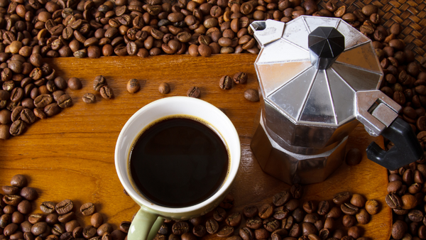 Auf einem Tisch steht ein alter Espressokocher und eine Tasse mit Kaffee. Daneben liegen viele Kaffeebohnen.