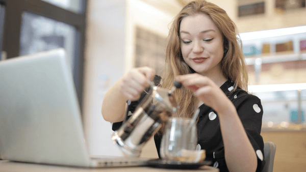Eine Frau sitzt vor ihrem Laptop und gießt sich gerade Kaffee aus der French Press in ihre Tasse ein