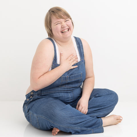 Kami wearing blue overalls sitting barefoot on the floor with her hand on her heart