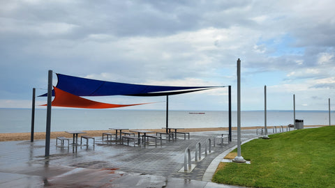 A beachfront seating area features blue and orange shade sails supported by sturdy sun shade posts, providing shelter from the sun for picnic tables below. The setup highlights the practical use of sun shade posts in creating a comfortable outdoor space by the water.