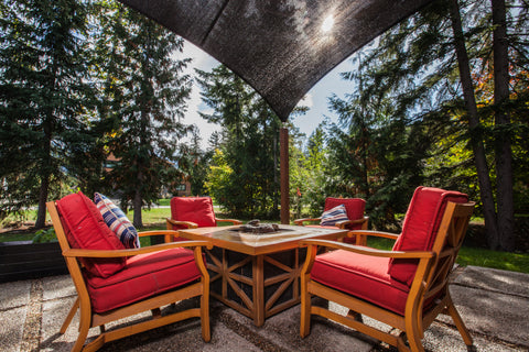 An outdoor patio area with wooden furniture featuring red cushions, arranged around a square fire pit table. The patio is shaded by a large black canopy attached to wooden posts. The backdrop includes lush green trees and a clear sky. This setup demonstrates how to attach a canopy to a house for a comfortable outdoor living space.