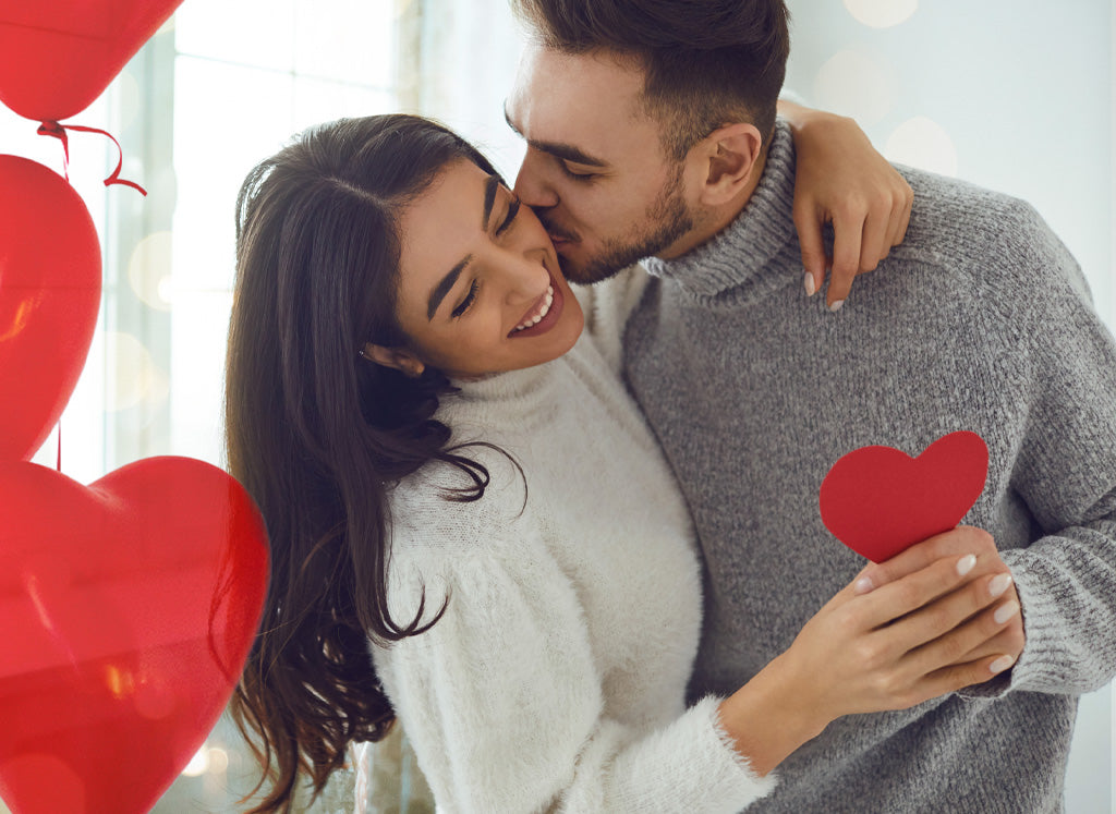 man kissing woman on cheek both holding a red heart shaped paper cutout