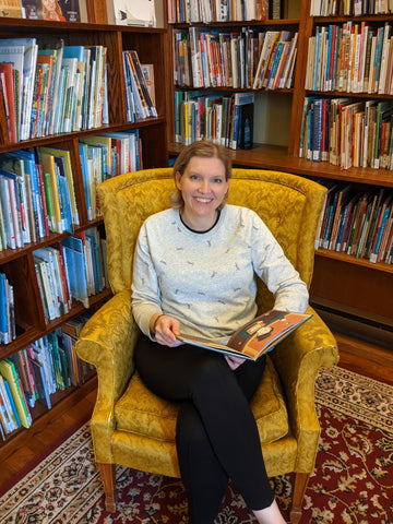 woman sitting in a yellow chair, an open book in her hand, surrounded by bookshelves