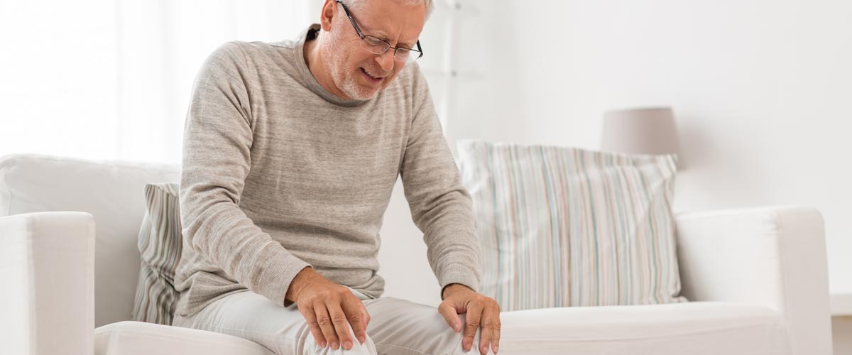 Älterer Mann hält sich die Beine und sitzt schmerzverzerrt auf der Couch. Er leidet an Durchblutungsstörungen