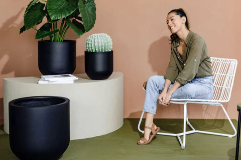 Happy woman seated beside elegant black Slugg planters with lush plants in a chic, minimalist indoor setting