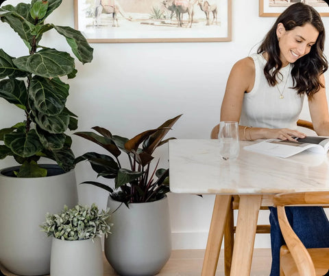 Smiling woman reading at a table with Slugg brand modern grey indoor planters with lush greenery in a bright room