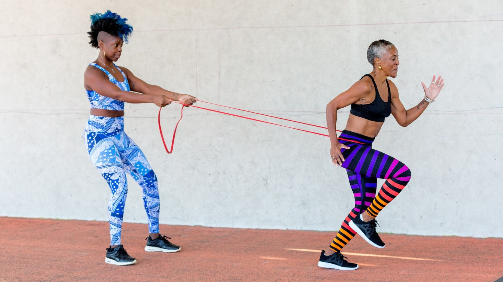 Women exercising. Woman on left is wearing a blue and white paisley bandana print top and bottom - print is called Imagine. Women on right is wearing black bra and striped pants in purple, pink, red, orange, and black - print is called Mabel Stripe.