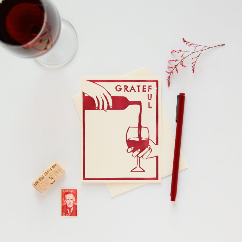 A letterpress printed thank you card made with hand-carved woodblocks on 100% post-consumer recycled paper with an image of a hand filling a wine glass from a bottle in a burgundy color that reads "grateful".