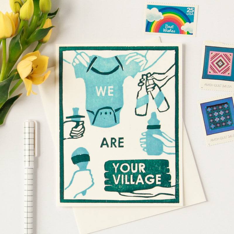 Greeting card for new babies and expecting parents that says "We Are Your Village" with a woodcut image of hands holding out a onesie, a baby bottle, a rattle, a pacifier, a pile of swaddle blankets and two bottles of beer in teal and forest green. 