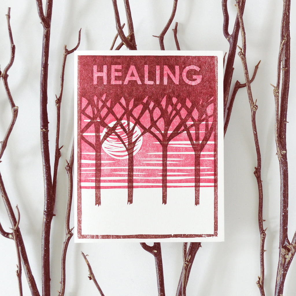 Handmade get well or feel better card that says "healing" with a woodcut image of a moon peaking through trees at sunrise in pink and maroon.