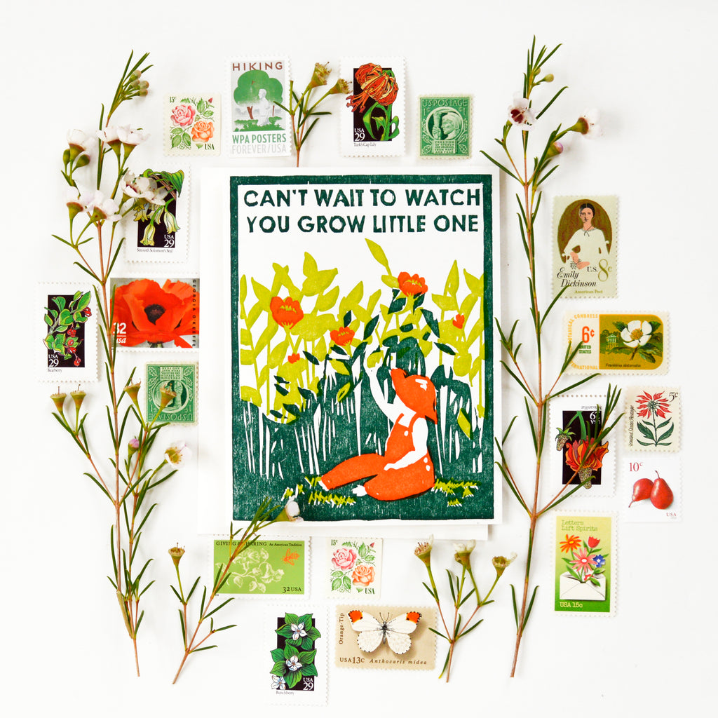 Handmade card for a new baby or new parents that says "can't wait to watch you grow little one" with a woodcut image of a child in red overalls and a red hat sitting in the grass in a garden, reaching for red flowers around her.