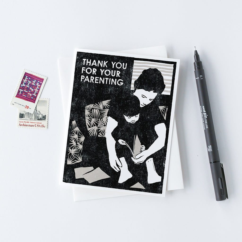 A mother's or father's day or thank you card with a black and silver woodcut image of a parent reading to a child on a couch.