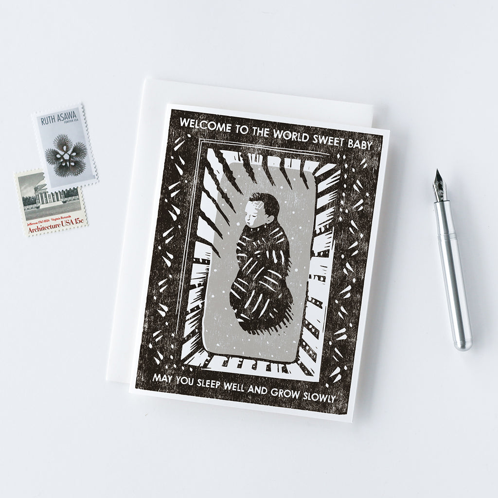 handmade new baby card with a woodcut image of a swaddled newborn asleep in a crib that says "Welcome to the World Sweet Baby, Sleep Well and Grow Slowly"