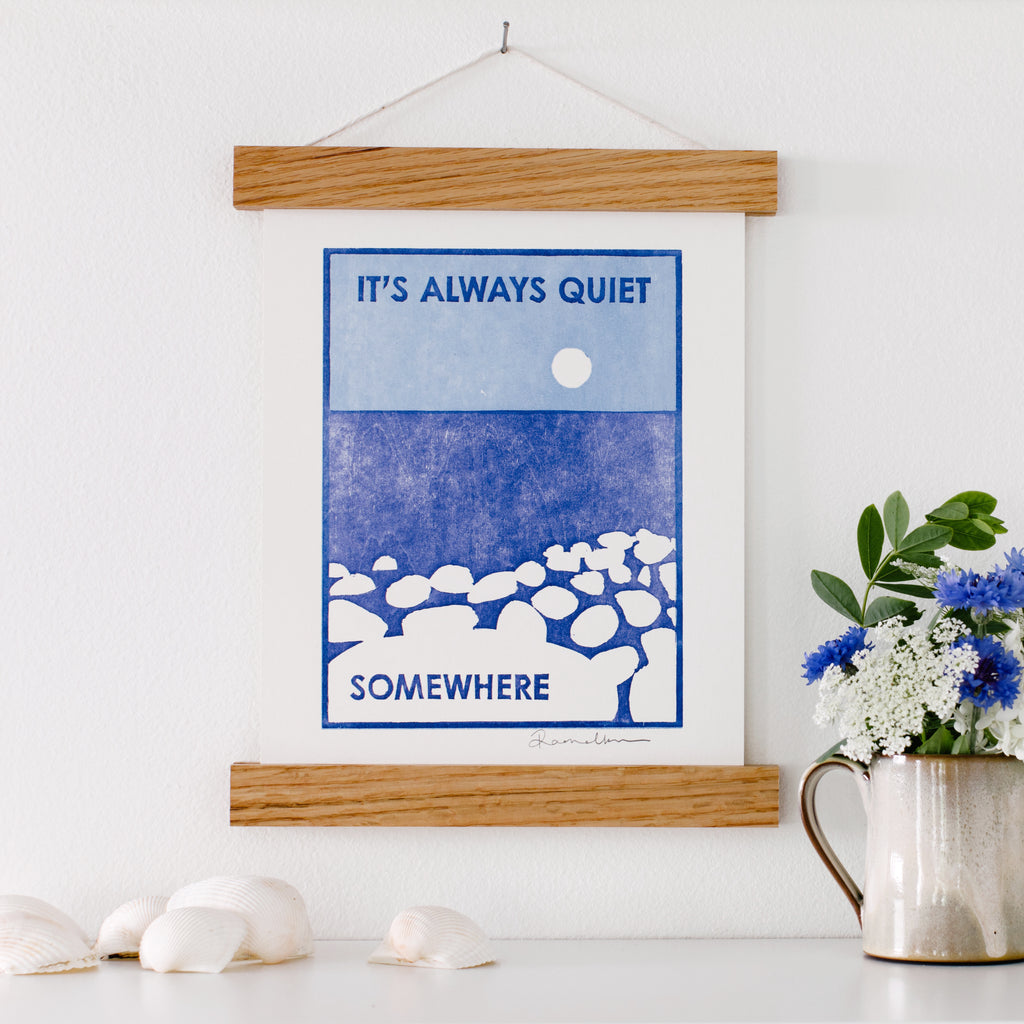 Letterpress woodcut art print of smooth stone beach on a blue lake with a white moon made with hand-carved woodblocks that says "It's Always Quiet Somewhere"