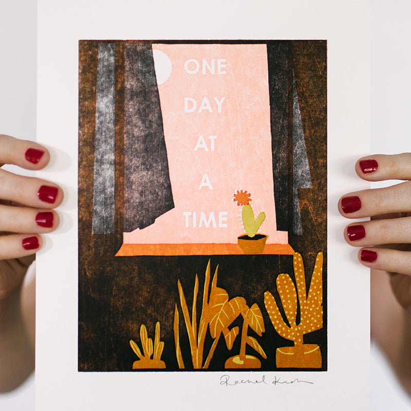 Letterpress woodcut art print of a cactus in a window with gauzy curtains and houseplants made with hand-carved woodblocks that says "One Day at a Time". 