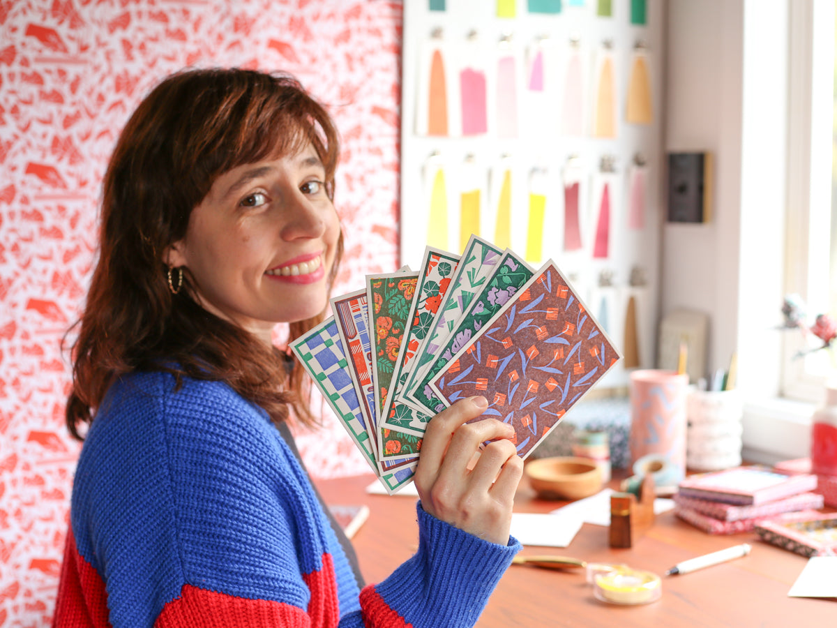 A photo of Heartell founder Rachel Kroh holding seven woodblock printed cards at her desk.