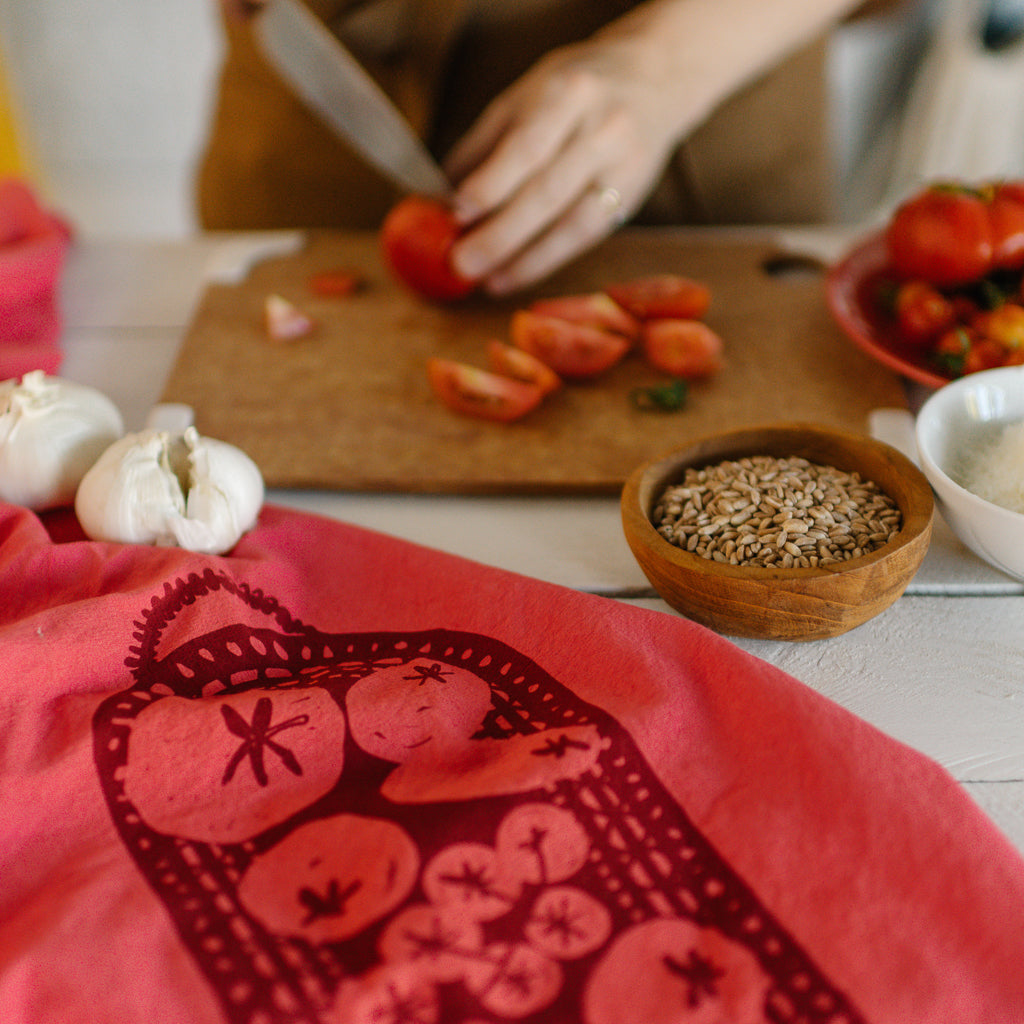 A woman slicing tomatoes with a red Generous Kitchen Towel with a screenprinted tomato design on it in the foreground.