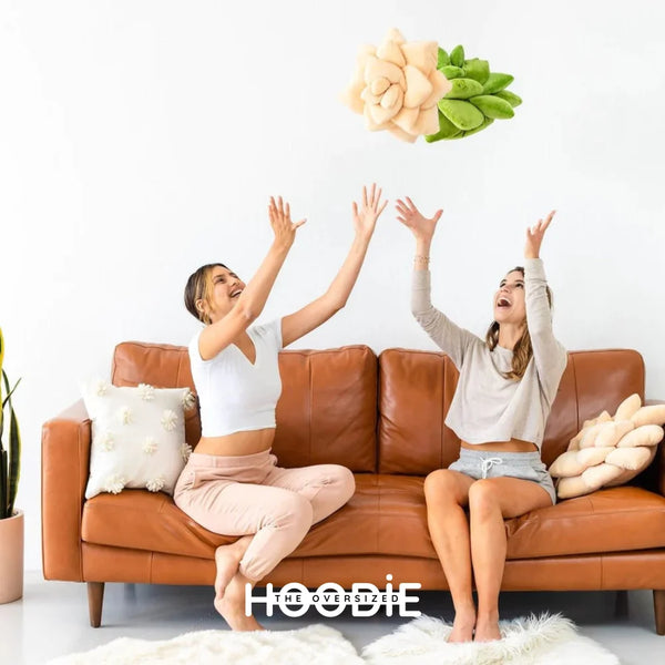 2 mujeres jóvenes en una sala de estar lanzando al aire 2 cojines de plantas Camélia