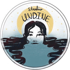 A drawing of a face in pale blue water with long hair floating in it. A yellow crescent crowns the head with black handwritten "studio UNDINE." The background is white with small yellow stars and is outlined with a circle.