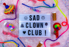 A photo of "Sad Clown Club !" in a custom text slider with emojis. The background is pink with chaotic strings, medication, tickets, and a spinning top toy.