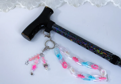 A photo of a black cane with sparkles on it and a pink, light blue, and white colored acrylic wrist strap.