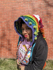 A rainbow colored long elf-style hat is worn by a black model. They are standing near a brick wall on grass. She is wearing a grey hoodie and a pink Mickie Mouse shirt