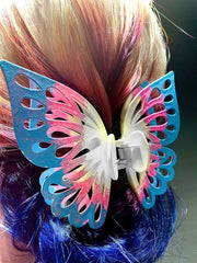 A butterfly hairpin that is painted light blue, light pink, and white like the trans flag. The hairpin is photographed in blonde hair with dark blue ends.