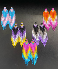 Multiple sets of Pride-themed earrings arranged in a V shape on a black table. Flag colors show the trans flag, nonbinary, pansexual, asexual, and lesbian. The beadwork on the earrings is a hexagonal shape with the striped colors in a V shape.