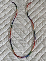 A rainbow colored necklace where the chain is made of red, orange, yellow, green, blue, and purple parts. The background is a white quilt