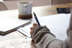 An image of a sweater-covered arm with a light skinned hand writing on paper. A coffee mug rests on the light wood desk in the background.