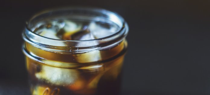 Image of cold brew coffee in a mason jar with ice. Mason jar in focus with depth-of-field against a dark background.