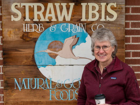 Lesa Wilson, owner and CFO of Caffe Ibis, standing in front of a historic Caffe Ibis sign with the text: "Straw Ibis Herb and Grain Co. - Natural & Good Foods""