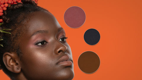 Black model with deep skin tone wearing all natural makeup by conscious beauty brand SAPPHO with three powder refills on a warm orange background