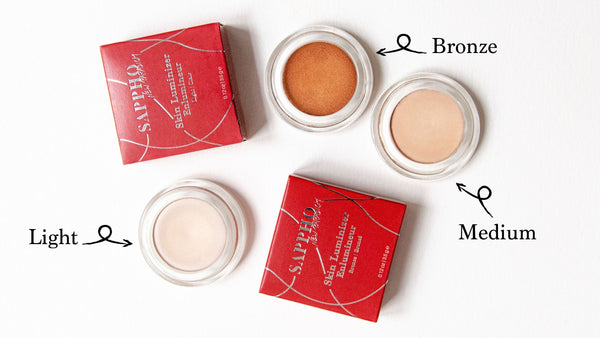 Three open skin luminizer pots showing shades Light, Medium & Bronze for all skin tones and two red recyclable box packaging