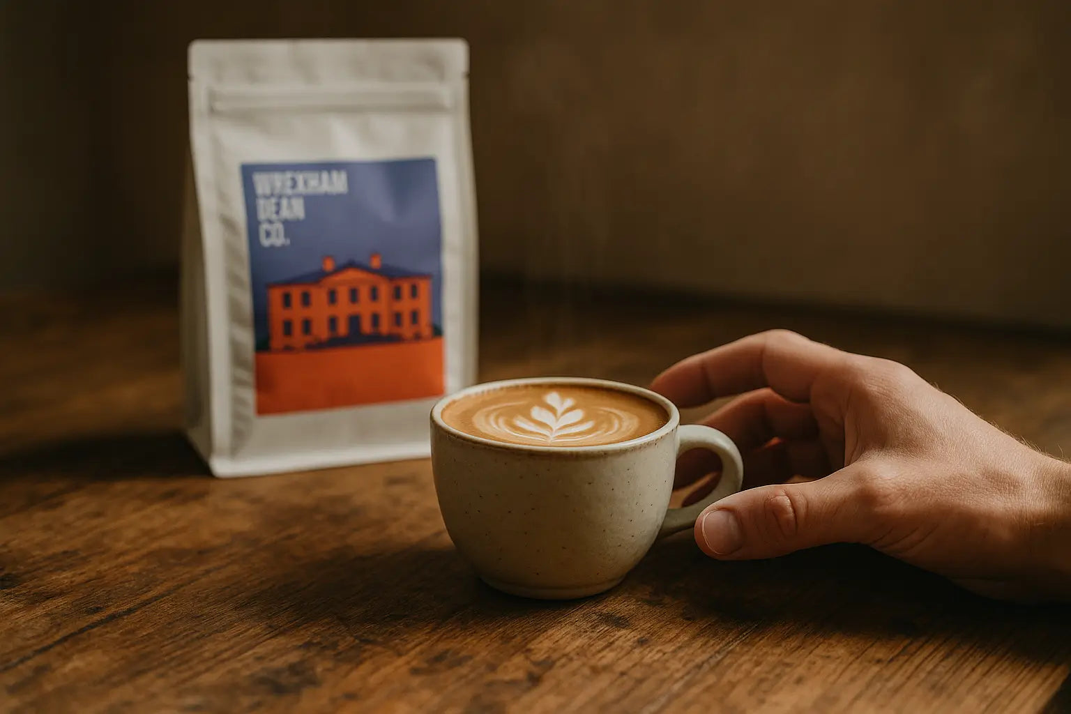 Flat white coffee in ceramic mug on rustic table, with hand reaching and Wrexham Bean Co. coffee bag blurred in the background