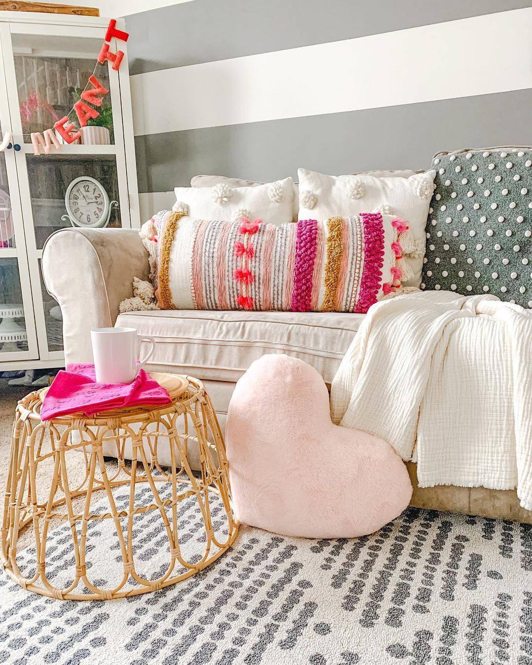 Geometric Arbor rug placed under a white couch. There are various pink and heart shaped pillows, a rattan ottoman and a garland hanging up behind the couch reading "be mine"
