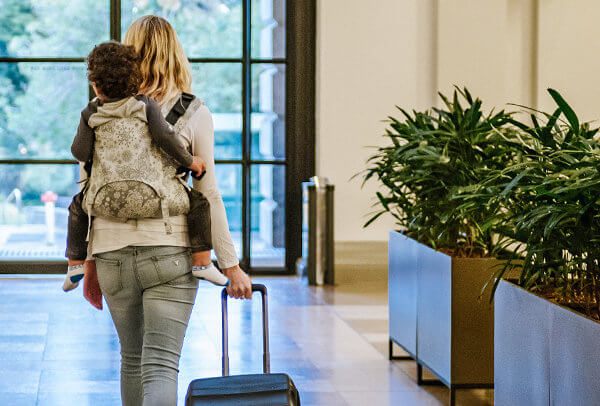 Mother carries toddler on her back and pushes a wheeled suitcase behind her