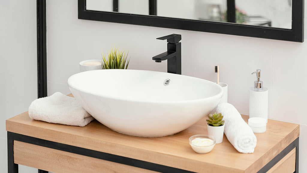 Modern bathroom with white sink on wooden table, black framed mirror, black faucet, potted plant, candle, soap dispenser, white bowl, and towel.